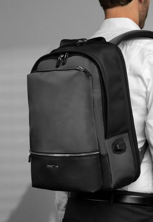 Vyras, dėvintis juodai pilką kuprinę su keliomis užtrauktukų skyriais ir USB įkrovimo lizdu šone, apsirengęs baltą marškinių ir juodus kelnes.