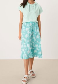 Haut court à capuche vert menthe clair associé à une jupe fluide longueur genou avec un motif de palmiers blancs. Sandales blanches épaisses.