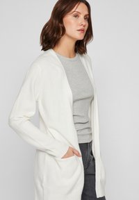 Cárdigan largo blanco hecho de un material de punto suave, con diseño de frente abierto, puños acanalados y bolsillos laterales, llevado sobre una camiseta gris.