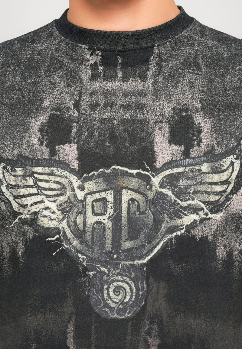 Close-up van een donkere, versleten T-shirt met een vervaagd gevleugeld embleem en de letters "RC" gecentreerd op de borst.