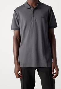 Polo gris avec un col, manches courtes et texture subtile. Il présente un petit logo noir sur la poitrine et des bords de manches rehaussés.