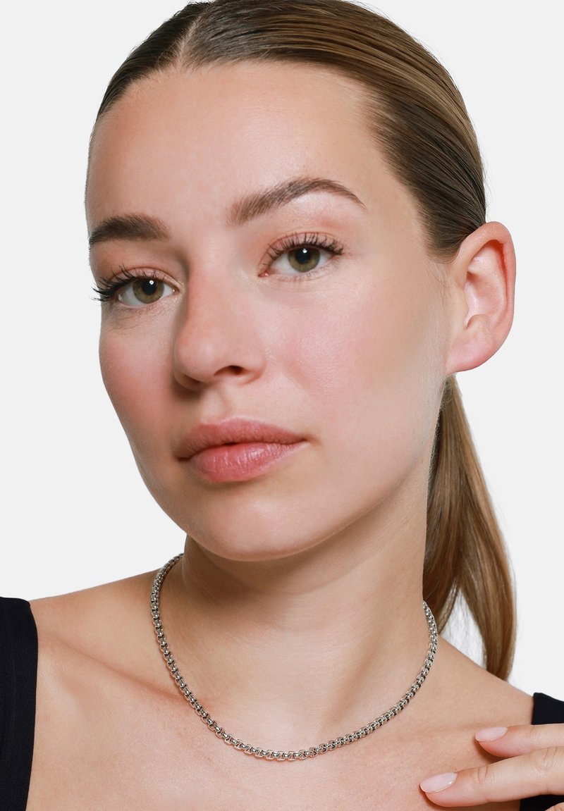 Sølvkjede med jevne ledd, hviler på en bar nakke, mot en hvit bakgrunn. Modellen har skulderlangt hår.