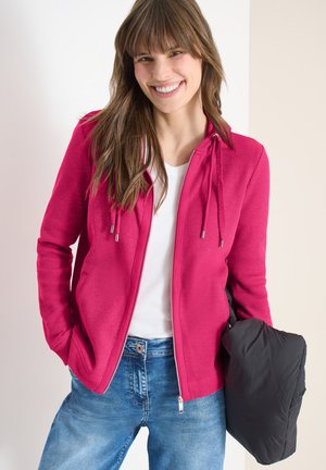 Femme souriante aux longs cheveux bruns portant une veste rose à fermeture éclair, un haut blanc, un jean bleu, et tenant un sac noir.