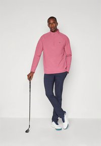 Pink polo stílusú pulóver félcipzárral, sötétkék nadrág, és fehér cipők kék díszítéssel, kezében golfütővel.