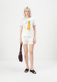 Hvit bomulls t-skjorte med et leopardmotiv og teksten "BAD VIBES", kombinert med hvite shorts og svarte sandaler. Mørk håndveske inkludert.