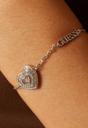 Zilveren schakelarmband met een hartvormige hanger versierd met kristallen en de merknaam "GUESS" om de pols.