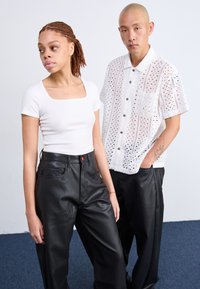 Zwarte leren broek, witte top met vierkante hals, en een witte gehaakte button-up shirt met een zak, met een textuurpatroon en korte mouwen.