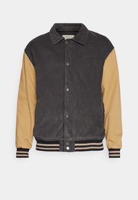 Veste en velours côtelé et tissu lisse avec un corps gris foncé et des manches beige. Elle présente des boutons-pression et des poignets et un ourlet à rayures côtelées.