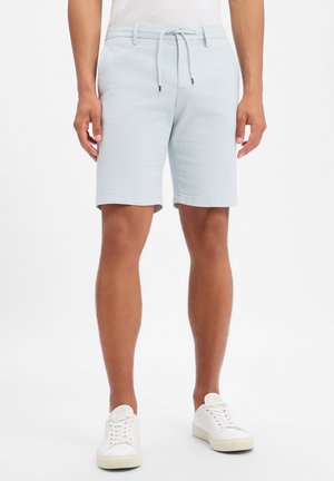 Mann trägt hellblaue Shorts mit Kordelzug und weiße Sneakers, steht vor einem schlichten weißen Hintergrund.