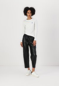 Camicia bianca a maniche lunghe con piccolo logo blu, abbinata a pantaloni in ecopelle neri e sneakers bianche con dettagli neri.