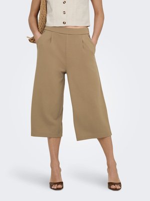 Kobieta ubrana w beżowe szerokie spodnie o długości do kostek, beżowy bezrękawnik zapinany na guziki, brązowe buty na obcasie z odkrytymi palcami, niesie plecioną torbę na ramieniu.