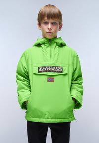 Ragazzo che indossa una giacca a vento verde brillante con tasca frontale etichettata "Napapijri" e una piccola bandiera norvegese, mani in tasca, sfondo neutro.
