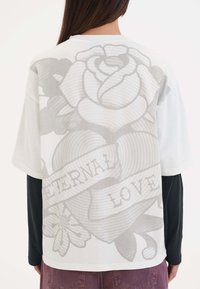 Witte oversized t-shirt met een grote grijze rozenafbeelding en de tekst "ETERNAL LOVE" op de achterkant, gemaakt van zachte stof en met een comfortabele pasvorm.