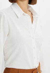 Mujer vestida con una camisa blanca de botones metida en unos pantalones marrones de cintura alta con un cinturón, mostrada de cuello a cintura.