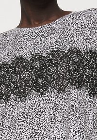 Černobílý top s tečkovaným vzorem a výrazným černým krajkovým akcentem přes hrudník. Kroužkový výstřih a volná tkanina.