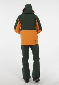 Technische Outdoorjacke in Grün und Orange, ausgestattet mit Kapuze, Reißverschlusstaschen und verstellbaren Bündchen. Isolierendes, wasserabweisendes Material.
