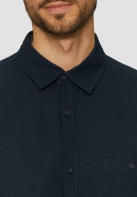 Camicia colletto a maniche corte di colore blu navy con tessuto strutturato, dotata di patta con bottoni e taschino sul petto. I bottoni sono di un blu navy scuro.