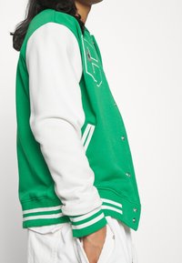 Chaqueta varsity verde y blanca con logotipo bordado, botones a presión, puños de rayas y paneles de hombro contrastantes. Textura de tela suave.