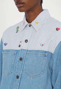 Denim és csíkos ing kék alappal, gombos kivitelezéssel, két első zsebbel és színes hímzett díszítésekkel a galléron.