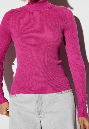Femme portant un pull à col roulé côtelé rose brillant et un pantalon gris clair avec des chaînes décoratives à la taille.