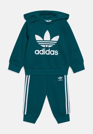 Completo in felpa Adidas color teal con cappuccio e pantaloni, caratterizzato da un grande logo bianco sul petto e strisce bianche lungo le gambe dei pantaloni. Tessuto morbido.