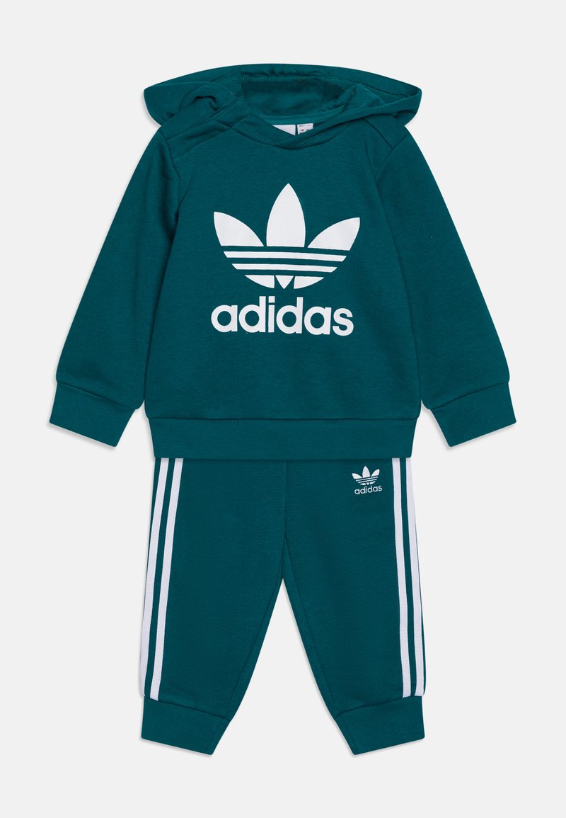Completo in felpa Adidas color teal con cappuccio e pantaloni, caratterizzato da un grande logo bianco sul petto e strisce bianche lungo le gambe dei pantaloni. Tessuto morbido.