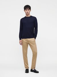 Mand står iført en mørkeblå langærmet skjorte, beige bukser og sorte pæne sko mod en ensartet lys baggrund.