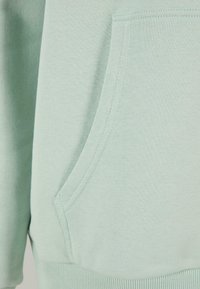 Tessuto felpato verde chiaro con una tasca frontale. La texture è morbida e le cuciture sono rifinite con cura. Orlo a coste nella parte inferiore.