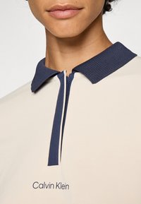 Beige polo shirt with a navy ribbed collar, featuring a front zipper and a small embroidered "Calvin Klein" logo on the chest.