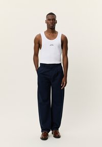 Λευκό αμάνικο μπλουζάκι με κεντημένο λογότυπο, συνδυασμένο με φαρδιά μπλε σκούρα παντελόνια. Το μοντέλο στέκεται με τα χέρια στις τσέπες, ολοκληρώνοντας το look με καφέ παπούτσια.