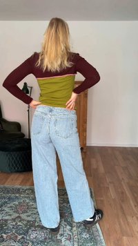 Femme aux cheveux blonds portant un haut à manches longues color block et un jean bleu clair ample, debout les mains sur les hanches dans un salon.