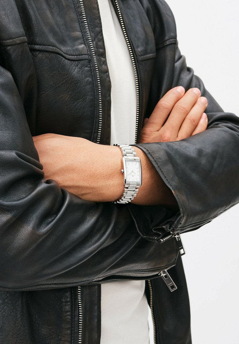 Zilveren rechthoekige horloge met een metalen schakeltband, gedragen over een zwart leren jack, met een gladde textuur en ritsaccenten.