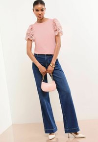 Haut rose clair avec des manches bouffantes en dentelle, associé à un jean en denim bleu à taille haute et jambes larges. Tenant un petit sac à main rose et portant des talons blancs.