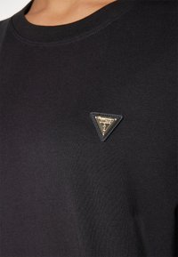 T-shirt nera in cotone con scollatura rotonda e un emblema triangolare dorato inserito sul lato sinistro. Tessuto liscio, design semplice.