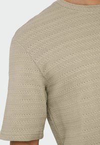 Lichtbeige gebreide trui met korte mouwen, voorzien van een textuurpatroon van horizontale lijnen en kleine openingen verspreid over de stof.