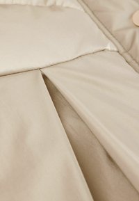 Giacca puffer beige con tessuto liscio e lucido e texture trapuntata; placca visibile e dettaglio in hardware arrotondato.