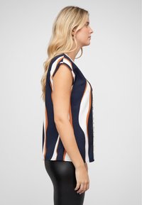 Haut bleu marine avec des rayures ondulées blanches et orange, à manches courtes et à encolure ronde. Tissu lisse, ourlet se terminant juste au-dessus des hanches.