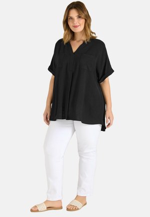 Vrouw die een losse zwarte blouse met korte mouwen, witte strakke broek en beige slippers draagt, staand tegen een effen achtergrond.