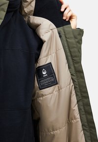 Gevoerde jas met een marineblauwe buitenlaag, beige voering en groene accenten. Beschikt over een label en een rits pocket aan de binnenkant. Zachte textuur.