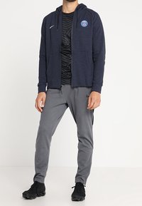 Sudadera navy con bolsillos frontales y logo, combinada con pantalones deportivos grises. Zapatillas negras completan el conjunto.