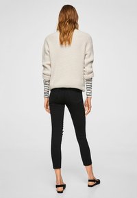Suéter de punto crema, jeans skinny negros con botones decorativos en el costado y sandalias planas negras. Camisa a rayas visible en los puños.