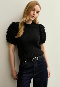 Maglione nero lavorato a maglia con maniche a sbuffo strutturate. Orlo a coste e colletto a girocollo. Indossato con jeans scuri a vita alta e una cintura nera con hardware argentato.