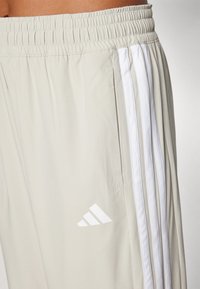 Helle beige Sporthosen mit elastischem Bund, drei weißen vertikalen Streifen an der Seite und einem weißen Logo in der Nähe des Saums.
