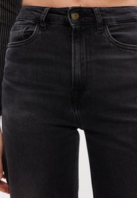 Črne jeans hlače z visokim pasom, ki imajo zlat gumb za zapiranje in subtilno teksturo. Stil s petimi žepi z temno strežbo.