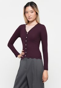 T-shirt bordeaux à manches longues avec décolleté en V, bord en dentelle et fermeture par boutons. Matériau texturé côtelé, associé à un pantalon taille haute gris.