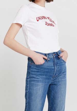 Personne portant un t-shirt blanc Calvin Klein Jeans rentré dans un jean en denim bleu taille haute, les mains dans les poches.
