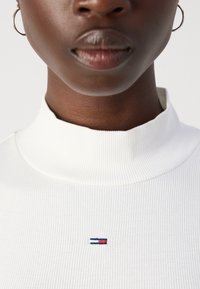 Baltos juostos megztinis su aukštu kaklu, kuriame yra nedidelis logotipas su mėlynomis ir raudonomis juostomis ant krūtinės. Audinys turi lygų, šiek tiek tekstūruotą išvaizdą.