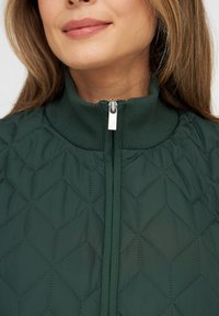 Grüne Steppjacke mit hohem Kragen, ausgestattet mit einem Reißverschluss vorne, sechseckigen Mustern und gerippten Akzenten am Halsauschnitt.