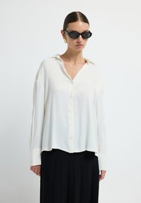 Chemisier blanc à boutons avec une coupe décontractée, en tissu légèrement transparent, et manches longues. Assorti à une jupe noire plissée et des lunettes de soleil.