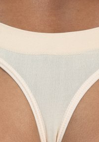 Beige String-Tanga aus einem glatten, dehnbaren Stoff. Mit einem breiten, flachen Gummibund und einem minimalistischen Design mit einem V-förmigen Schnitt.